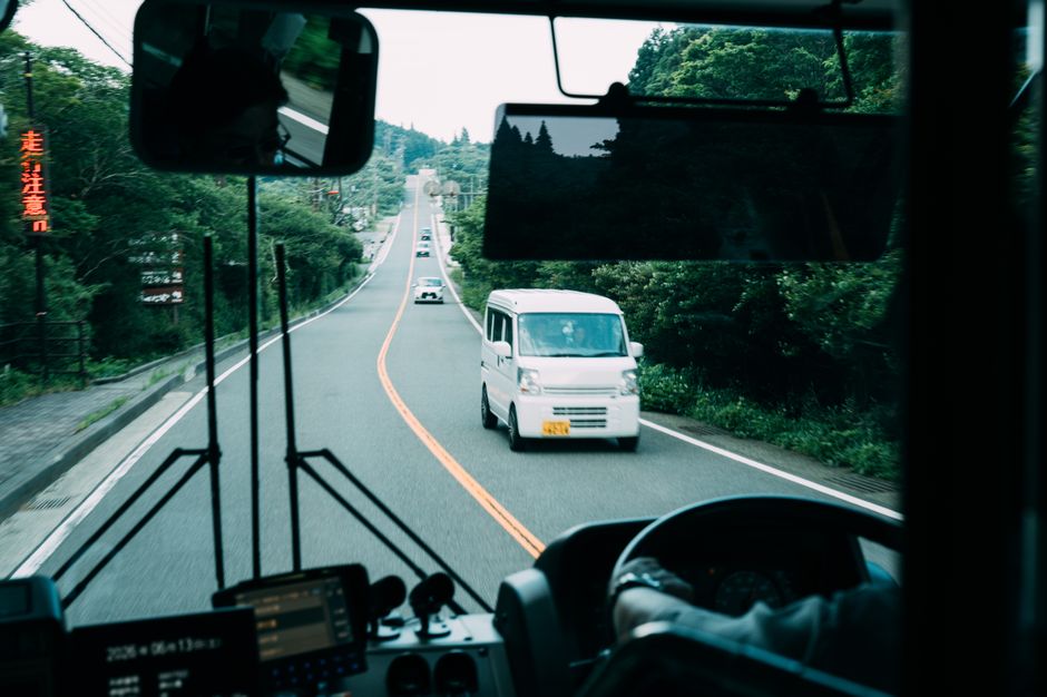 緑豊かな景色の良い道を走るバスの車内から見た景色。白いバンが通り過ぎていく。